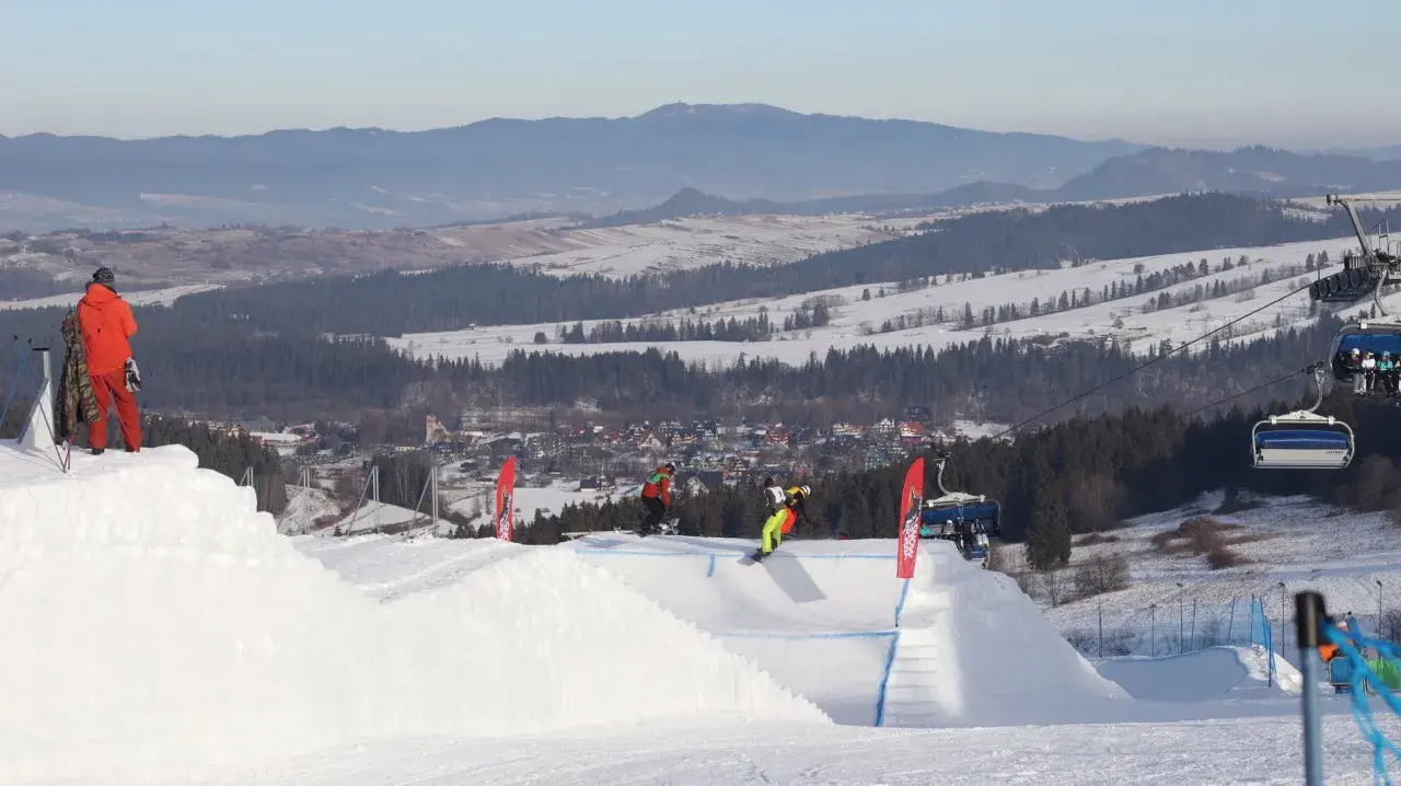 Snowboardziści na stoku z wyciągiem krzesełkowym i widokiem na góry i miasteczko.