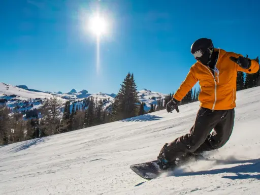 Snowboardzista w pomarańczowej kurtce zjeżdża po ośnieżonym stoku w słoneczny dzień.