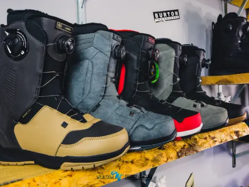Rząd butów snowboardowych w różnych kolorach i stylach, ustawionych na drewnianej półce.