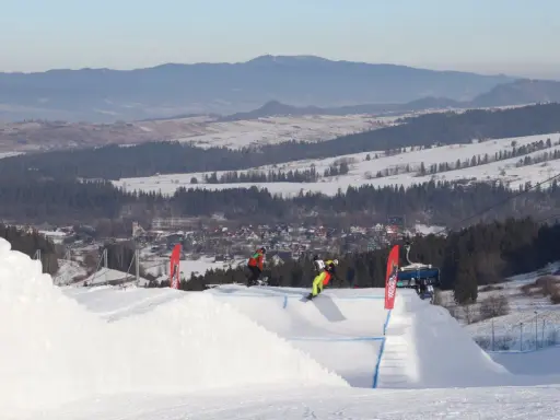 Snowboardziści na stoku z wyciągiem krzesełkowym i widokiem na góry i miasteczko.