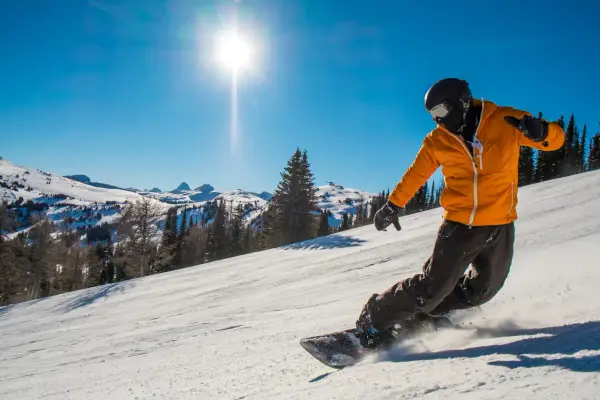 Snowboardzista w pomarańczowej kurtce zjeżdża po ośnieżonym stoku w słoneczny dzień.